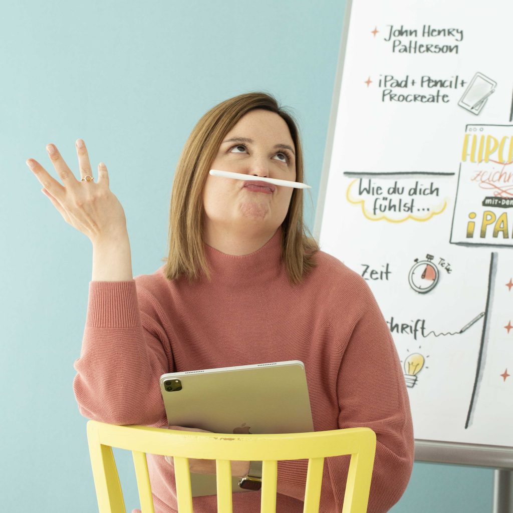 Sylvia sitzt auf einem Stuhl vor einem Flipchart. Das iPad im Arm, der ApplePen ist zwischen Oberlippe und Nase geklemmt. Sylvia hat einen rätselnden Blick nach oben.