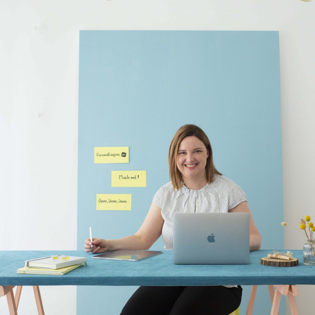 Sylvia am Arbeitstisch mit iPad und Laptop und Blick in die Kamera.